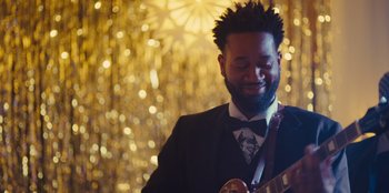 Movie still from “Stargirl” (2020), directed by Julia Hart – A man in a suit and bow tie playing a guitar in front of lights; Close Up shot, Over the shoulder angle