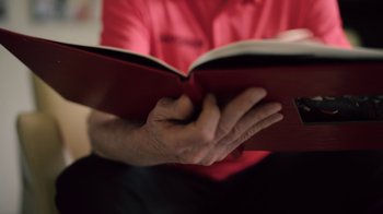 Movie still from “Stephen Curry: Underrated” (2023), directed by Peter Nicks – An older man reading a book in a red shirt; Close Up shot, High angle