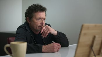 Movie still from “Still: A Michael J. Fox Movie” (2023), directed by Davis Guggenheim – A man sitting at a table with his hand under his chin; Close Up shot, Over the shoulder angle