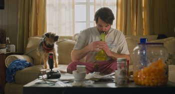 Movie still from “Strays” (2023), directed by Josh Greenbaum – A man sitting on a couch next to a dog; Medium shot, Low angle