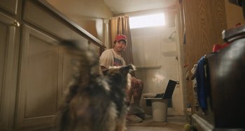 Movie still from “Strays” (2023), directed by Josh Greenbaum – A man sitting in a bathroom next to a dog; Wide shot, Low angle