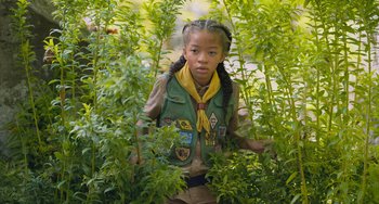 Movie still from “Strays” (2023), directed by Josh Greenbaum – A girl scout is surrounded by plants in a forest; Medium shot, High angle