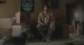 Movie still from “Strays” (2023), directed by Josh Greenbaum – A man sitting on a chair in a living room; Medium shot, Low angle