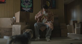 Movie still from “Strays” (2023), directed by Josh Greenbaum – A man sitting on a chair in a living room; Wide shot, Low angle