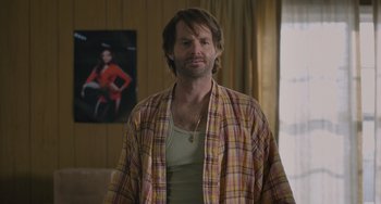 Movie still from “Strays” (2023), directed by Josh Greenbaum – A man in a plaid shirt standing in front of a picture; Medium shot, Over the shoulder angle