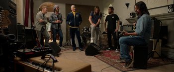 Movie still from “Studio 666” (2022), directed by BJ McDonnell – A group of men standing around a microphone; Wide shot, High angle