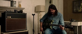 Movie still from “Studio 666” (2022), directed by BJ McDonnell – A man sitting down playing an electric guitar in a room; Medium shot, Over the shoulder angle