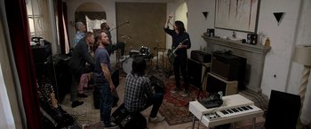 Movie still from “Studio 666” (2022), directed by BJ McDonnell – A group of people in a living room playing instruments; Wide shot, High angle