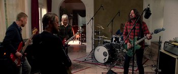 Movie still from “Studio 666” (2022), directed by BJ McDonnell – A group of people that are in a room with instruments; Wide shot, Over the shoulder angle