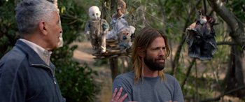 Movie still from “Studio 666” (2022), directed by BJ McDonnell – A man with long hair standing next to a tree with dolls hanging from it; Medium shot, Over the shoulder angle