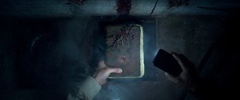 Movie still from “Studio 666” (2022), directed by BJ McDonnell – A person holding a book in a room with smoke coming out of it; Extreme Close Up shot, Overhead angle
