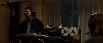 Movie still from “Studio 666” (2022), directed by BJ McDonnell – A man with headphones playing drums in a room; Medium shot, Over the shoulder angle