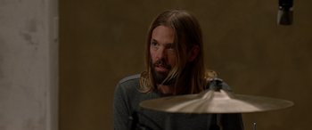 Movie still from “Studio 666” (2022), directed by BJ McDonnell – A man with long hair and a beard is sitting next to a cymbal; Close Up shot, Over the shoulder angle