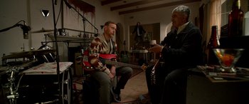 Movie still from “Studio 666” (2022), directed by BJ McDonnell – A man sitting on a chair holding a guitar; Wide shot, Over the shoulder angle