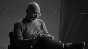 Movie still from “Stutz” (2022), directed by Jonah Hill – An older man sitting in a chair writing on a notebook; Medium shot, Low angle