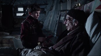 Movie still from “Charlie and the Chocolate Factory” (2005), directed by Tim Burton – An older man sitting next to a young boy holding a pair of knifes; Medium shot, Over the shoulder angle