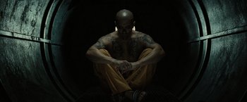 Movie still from “Suicide Squad” (2016), directed by David Ayer – A man sitting on the ground with his arms crossed; Medium shot, High angle