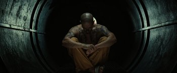 Movie still from “Suicide Squad” (2016), directed by David Ayer – A man sitting on the ground with his hands crossed; Medium shot, High angle