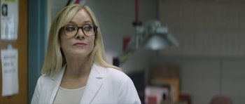 Movie still from “Suitable Flesh” (2023), directed by Joe Lynch – A woman wearing a lab coat and glasses in a room; Close Up shot, Over the shoulder angle