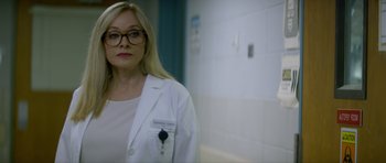 Movie still from “Suitable Flesh” (2023), directed by Joe Lynch – A woman in a white lab coat standing in front of a wall; Close Up shot, Over the shoulder angle