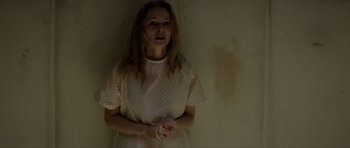 Movie still from “Suitable Flesh” (2023), directed by Joe Lynch – A woman standing in a room with her hands clasped together; Medium shot, Over the shoulder angle