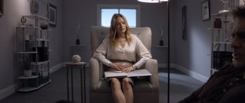 Movie still from “Suitable Flesh” (2023), directed by Joe Lynch – A woman is sitting in a chair reading a book; Medium shot, Over the shoulder angle