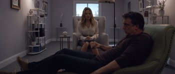 Movie still from “Suitable Flesh” (2023), directed by Joe Lynch – A woman sitting in a chair in front of a man; Wide shot, Over the shoulder angle
