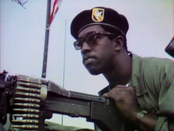 Movie still from “Summer of Soul (...Or, When the Revolution Could Not Be Televised)” (2021), directed by Questlove – A man in a hat and glasses is holding a gun; Close Up shot, Low angle