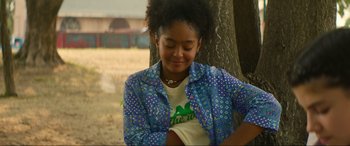 Movie still from “Summering” (2022), directed by James Ponsoldt – A young girl sitting in front of a tree smiling; Medium shot, Over the shoulder angle