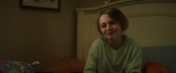 Movie still from “Summering” (2022), directed by James Ponsoldt – A young girl sitting in a room smiling for the camera; Close Up shot, Over the shoulder angle