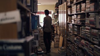 Movie still from “Summertime” (2020), directed by Carlos López Estrada – A man standing in a room filled with lots of books; Medium shot, Over the shoulder angle