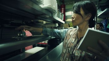Movie still from “Summertime” (2020), directed by Carlos López Estrada – A woman in a kitchen reaching for a stack of food; Medium shot, Low angle