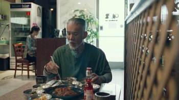 Movie still from “Summertime” (2020), directed by Carlos López Estrada – An older man sitting at a table eating food; Medium shot, Over the shoulder angle