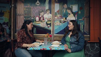 Movie still from “Summertime” (2020), directed by Carlos López Estrada – Two women sitting at a table with food in front of a painting; Wide shot, Low angle