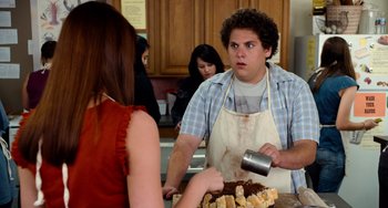 Movie still from “Superbad” (2007), directed by Greg Mottola – A man in an apron is holding a pan of cake while a woman in an apron watches; Medium shot, Over the shoulder angle
