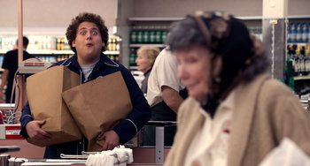 Movie still from “Superbad” (2007), directed by Greg Mottola – A man holding a brown paper bag in front of a woman; Medium shot, Over the shoulder angle