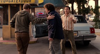 Movie still from “Superbad” (2007), directed by Greg Mottola – A man being held back by another man and another man standing in front of a car; Medium shot, Over the shoulder angle