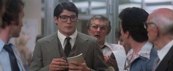 Movie still from “Superman” (1978), directed by Richard Donner – A man in a suit and glasses is holding a book; Medium shot, Over the shoulder angle