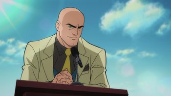 Movie still from “Superman: Man of Tomorrow” (2020), directed by Chris Palmer – A bald man in a suit and tie is sitting at a podium; Medium shot, Low angle