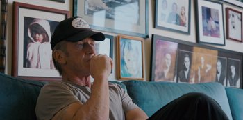 Movie still from “Superpower” (2023), directed by Aaron Kaufman – A man sitting on a couch in front of a wall of pictures; Close Up shot, Over the shoulder angle