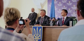 Movie still from “Superpower” (2023), directed by Aaron Kaufman – A man in a suit and tie sitting at a podium; Medium shot, Low angle