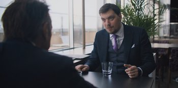 Movie still from “Superpower” (2023), directed by Aaron Kaufman – A man sitting at a table in front of a glass of water; Medium shot, Over the shoulder angle