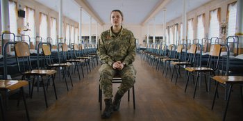 Movie still from “Superpower” (2023), directed by Aaron Kaufman – A soldier sitting on top of a chair in front of rows of chairs; Wide shot, Low angle