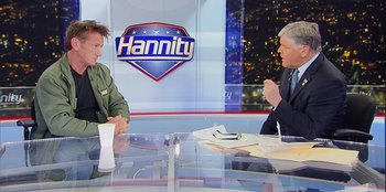 Movie still from “Superpower” (2023), directed by Aaron Kaufman – Two people sitting at a table in front of a hannity sign; Medium shot, Over the shoulder angle
