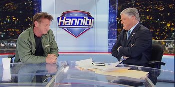 Movie still from “Superpower” (2023), directed by Aaron Kaufman – Two people sitting at a table in front of a sign; Medium shot, Over the shoulder angle