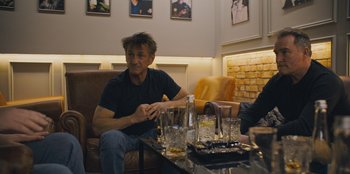 Movie still from “Superpower” (2023), directed by Aaron Kaufman – A man sitting on a couch in front of a coffee table; Medium shot, Over the shoulder angle