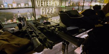 Movie still from “Superpower” (2023), directed by Aaron Kaufman – Several guns are laying on a table in front of a sign that reads " veterano pizza "; Extreme Close Up shot, High angle