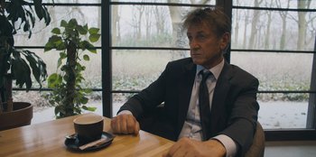 Movie still from “Superpower” (2023), directed by Aaron Kaufman – A man sitting at a wooden table in front of a window; Medium shot, Over the shoulder angle