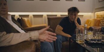 Movie still from “Superpower” (2023), directed by Aaron Kaufman – A man sitting on a brown leather couch next to another man; Medium shot, Over the shoulder angle