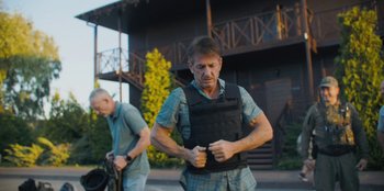 Movie still from “Superpower” (2023), directed by Aaron Kaufman – A man wearing a body armor while standing next to another man; Medium shot, Over the shoulder angle